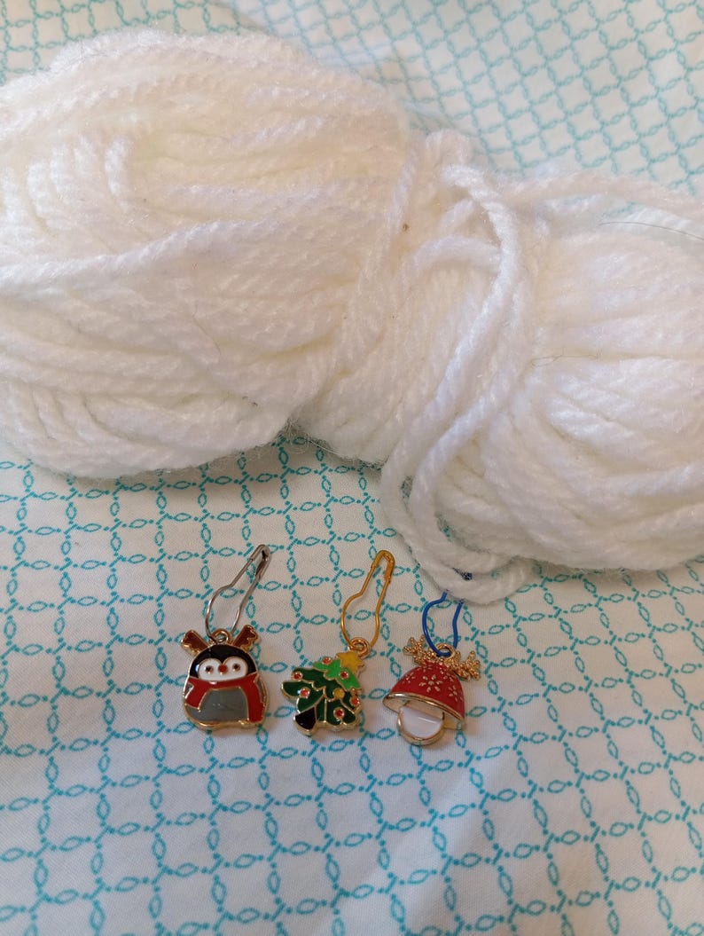 May include: Three Christmas themed stitch markers with a penguin, a Christmas tree, and a bell. The markers are gold with colorful enamel details. The markers are on a white background.