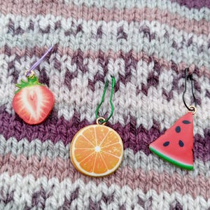 May include: Three fruit-themed stitch markers: a strawberry, an orange slice, and a watermelon slice. Each marker has a colorful fruit design and a metal loop. The markers are on a textured, multi-colored knitted background.