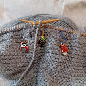 May include: Gray knitted fabric with three Christmas-themed stitch markers. The markers are a penguin, a Christmas tree, and a bell.
