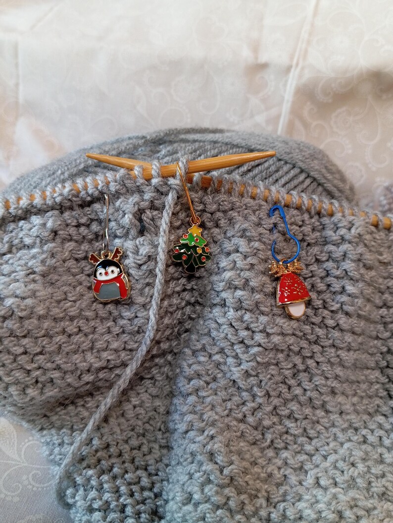 May include: Gray knitted fabric with three Christmas-themed stitch markers. The markers are a penguin, a Christmas tree, and a bell.