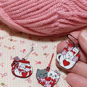 May include: Three enamel pin stitch markers featuring cats and hearts. The first pin is a jar with a red lid and the text "Full of Love" on the label. The second pin is a heart with two cats inside and pink and red hearts. The third pin is a white mug with a red heart and two cats inside.