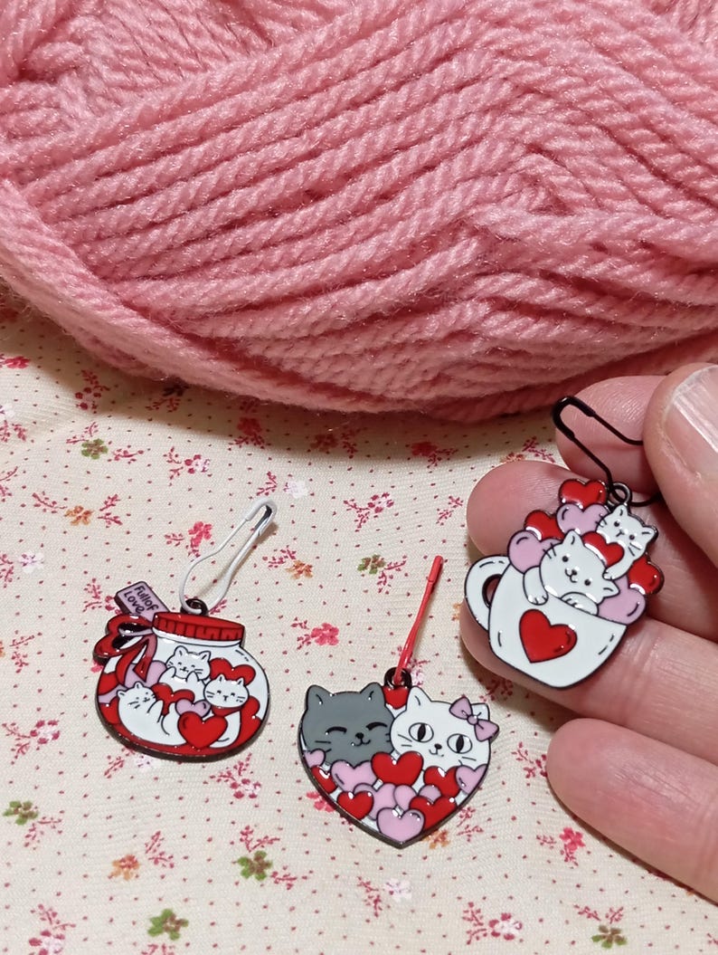 May include: Three enamel pin stitch markers featuring cats and hearts. The first pin is a jar with a red lid and the text "Full of Love" on the label. The second pin is a heart with two cats inside and pink and red hearts. The third pin is a white mug with a red heart and two cats inside.