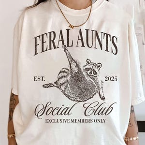 May include: White t-shirt with a black graphic of a raccoon with the text "Feral Aunts Social Club" and "Est. 2025" printed on it.