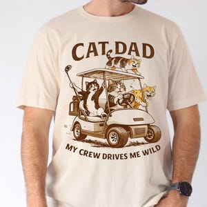 Puede incluir: Camiseta beige con un gráfico de gatos en un carrito de golf. El texto "CAT DAD" está encima del carrito, y "MY CREW DRIVES ME WILD" debajo. El carrito de golf tiene cuatro gatos, y uno de ellos sostiene un palo de golf.