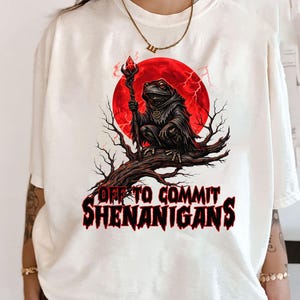 May include: Cream-colored t-shirt featuring a graphic of a frog in a hooded cloak holding a staff, set against a red moon and tree branches. The text "OFF TO COMMIT SHENANIGANS" is printed below the image in red.
