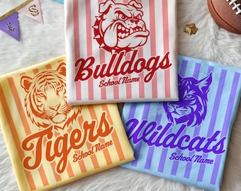 Może przedstawiać: Trzy złożone koszulki w pionowe paski. Biała koszulka ma czerwoną grafikę buldoga i napis Bulldogs School Name. Żółta koszulka ma pomarańczową grafikę tygrysa i napis Tigers School Name. Niebieska koszulka ma fioletową grafikę dzikiego kota i napis Wildcats School Name.
