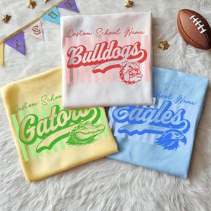 Peut inclure: Trois t-shirts avec des noms d'école personnalisés et des graphismes de mascotte. Le t-shirt blanc affiche Bulldogs, le jaune montre Gators et le bleu clair présente Eagles. Chaque t-shirt a des rayures verticales et une illustration.