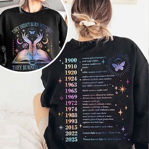 May include: Black sweatshirt with the text "THEY DIDN'T BURN WITCHES THEY BURNED WOMEN" and a graphic of an open book, hands, and a butterfly. The back of the sweatshirt lists women's rights milestones from 1900 to 2025.