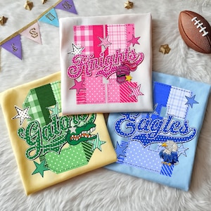 May include: Three folded t-shirts featuring large block letters with patchwork patterns and sports team graphics. The white shirt displays Knights with a helmet, the yellow shirt shows Gators with an alligator, and the blue shirt features Eagles with an eagle.