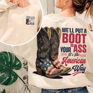 May include: Cream-colored sweatshirt with a graphic of cowboy boots featuring the American flag and the text "WE'LL PUT A BOOT IN YOUR ASS It's The American Way." A smaller graphic on the front says "BROUGHT TO YOU COURTESY OF RED, WHITE & BLUE."