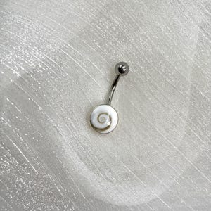 Puede incluir: Un piercing para el ombligo plateado con una barra curva y una pequeña bola en la parte superior. El dije inferior es un disco blanco con un diseño en espiral, sobre un fondo blanco.