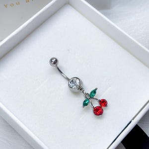 Puede incluir: Un piercing para el ombligo plateado con una barra curva. Tiene una gema transparente y un dije de dos gemas rojas en forma de cereza con hojas verdes. La joya se presenta en una caja blanca.
