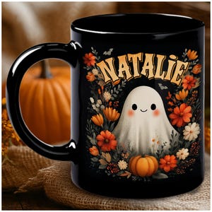 Op de afbeelding: Zwarte keramische mok met een Halloween-ontwerp. Het heeft een illustratie van een lachend spook omringd door oranje en witte bloemen en een kleine pompoen. De naam "Natalie" staat in oranje boven het spook.