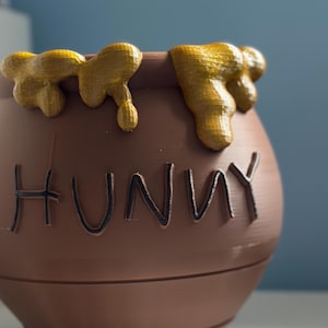 May include: A brown ceramic honey pot with the word "Hunny" written on the front. The pot is decorated with yellow honey dripping down the sides.