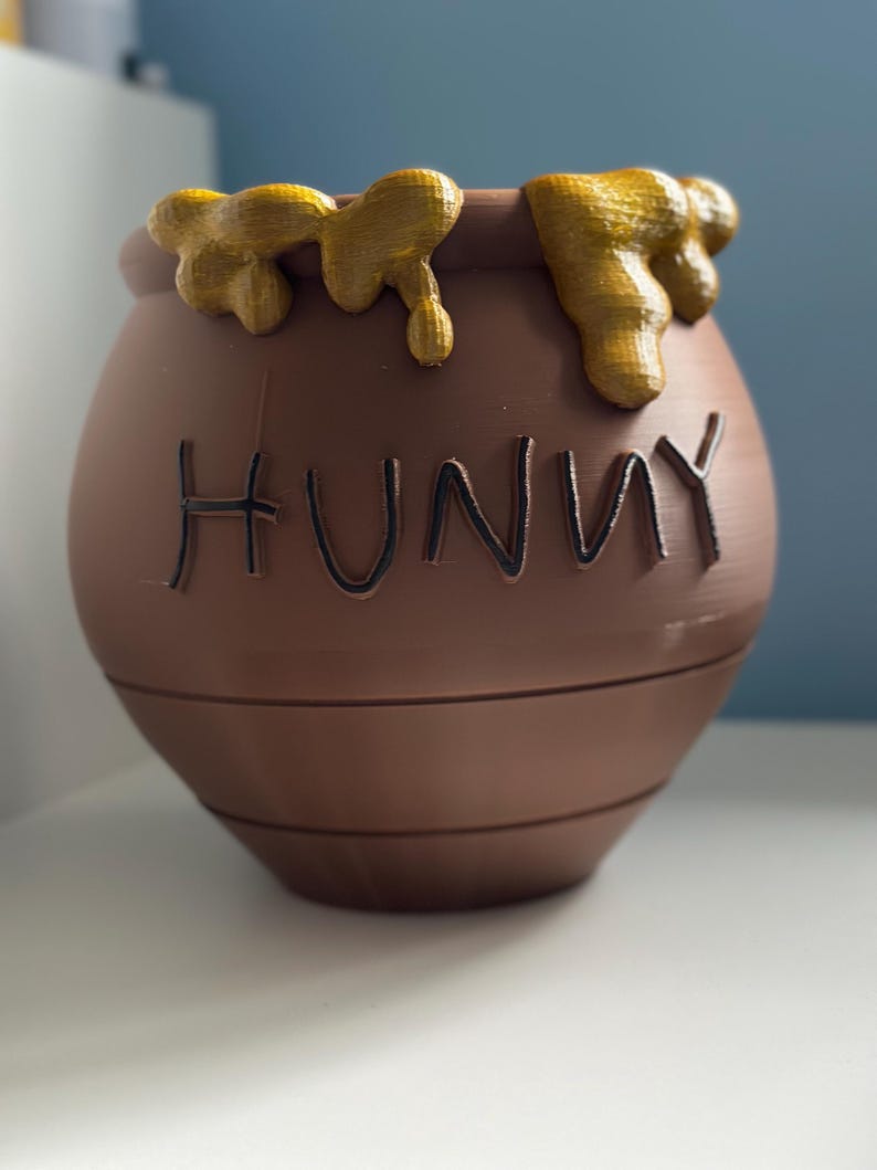 May include: A brown ceramic honey pot with the word "Hunny" written on the front. The pot is decorated with yellow honey dripping down the sides.