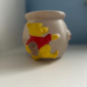 Puede incluir: Un tarro de miel de cerámica de Winnie the Pooh con un diseño de Winnie the Pooh amarillo y rojo.