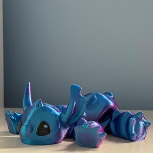 May include: A blue and purple iridescent 3D printed figurine of Stitch from Lilo & Stitch. The figurine is lying on its belly with its arms and legs outstretched.