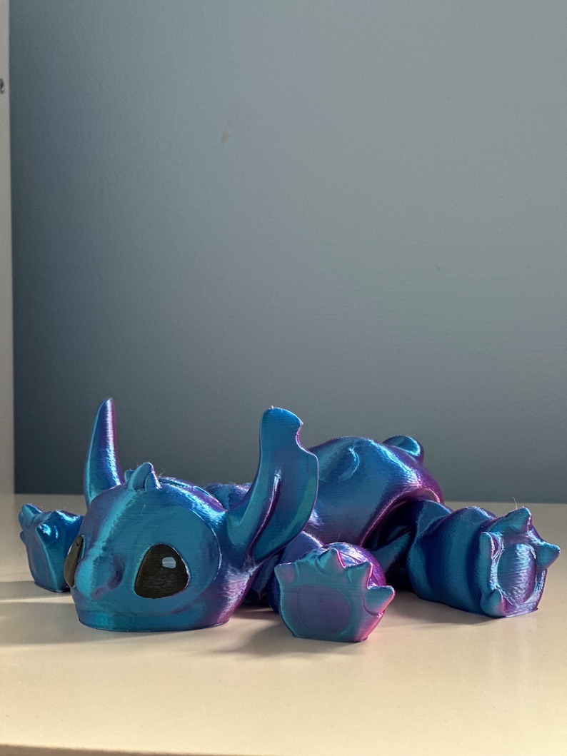 May include: A blue and purple iridescent 3D printed figurine of Stitch from Lilo & Stitch. The figurine is lying on its belly with its arms and legs outstretched.