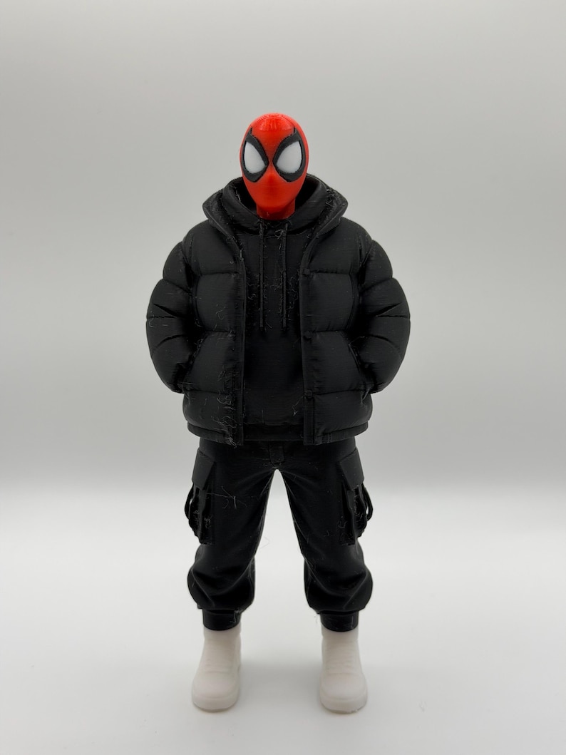 May include: A collectible action figure featuring a red mask with large white eye lenses, wearing a black hooded puffer jacket over a black hoodie, black cargo pants with side pockets, and plain white boots, standing in a neutral pose.