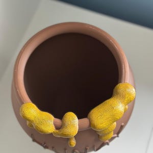 May include: A brown ceramic honey pot with a yellow honey drip design. The pot is empty and has the word "Hunny" written on the side.