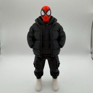 May include: A collectible action figure featuring a red mask with large white eye lenses, wearing a black hooded puffer jacket over a black hoodie, black cargo pants with side pockets, and plain white sneakers, standing in a neutral pose.
