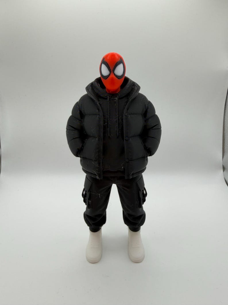 May include: A collectible action figure featuring a red mask with large white eye lenses, wearing a black hooded puffer jacket over a black hoodie, black cargo pants with side pockets, and plain white sneakers, standing in a neutral pose.
