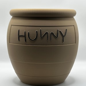 May include: A brown, barrel-shaped container with the word "HUNNY" in black lettering. The container has a ridged top and horizontal bands around the body. The background is plain white.