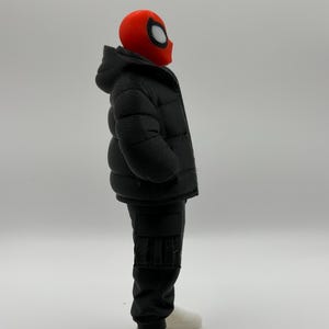 May include: A figurine featuring a red mask with white eye lenses, wearing a black hooded puffer jacket, black cargo pants, and white sneakers. The character is shown in profile, standing upright with a neutral posture against a plain background.