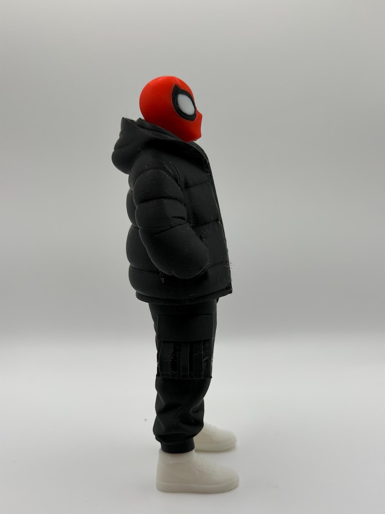 May include: A figurine featuring a red mask with white eye lenses, wearing a black hooded puffer jacket, black cargo pants, and white sneakers. The character is shown in profile, standing upright with a neutral posture against a plain background.