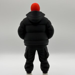 May include: A figurine of a person viewed from the back, wearing a bright red beanie, a black hooded puffer jacket with horizontal quilted sections, black cargo pants with side pockets, and plain white shoes, standing against a neutral background.