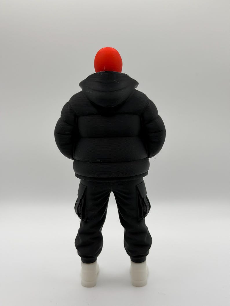 May include: A figurine of a person viewed from the back, wearing a bright red beanie, a black hooded puffer jacket with horizontal quilted sections, black cargo pants with side pockets, and plain white shoes, standing against a neutral background.