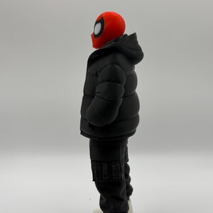 May include: A figurine featuring a red mask with large white eye lenses, wearing a black hooded puffer jacket, black cargo pants with side pockets, and white sneakers. The figure is shown from a side profile view against a plain background.