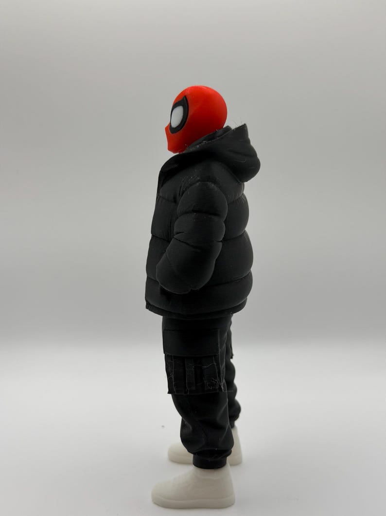 May include: A figurine featuring a red mask with large white eye lenses, wearing a black hooded puffer jacket, black cargo pants with side pockets, and white sneakers. The figure is shown from a side profile view against a plain background.