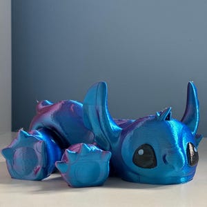 May include: A blue and purple iridescent 3D printed figurine of Stitch from Lilo & Stitch. The figurine is lying on its side with its head resting on the ground. Stitch has large, black eyes and a wide, toothy grin.