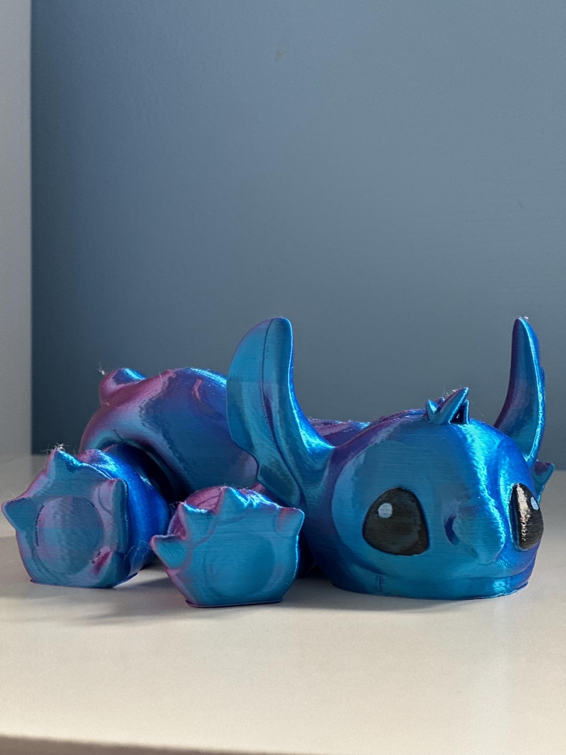 May include: A blue and purple iridescent 3D printed figurine of Stitch from Lilo & Stitch. The figurine is lying on its side with its head resting on the ground. Stitch has large, black eyes and a wide, toothy grin.