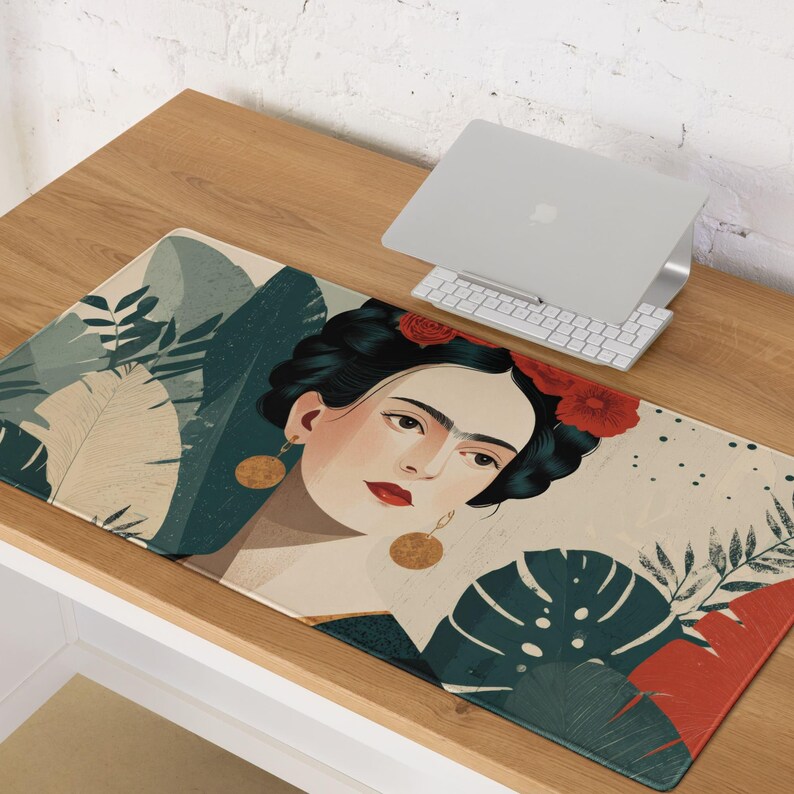May include: A rectangular desk mat featuring a portrait of a woman with dark hair, red flowers, and gold earrings. The mat has a beige background with green and red foliage. A laptop and keyboard are on the wooden desk.