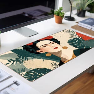 May include: Rectangular desk mat featuring a stylized portrait of a woman with dark hair and red flowers, surrounded by green and beige tropical foliage. The mat is on a white desk, with a computer and plants in the background.