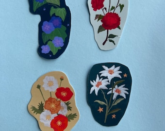 Può includere: Quattro adesivi fustellati con illustrazioni botaniche. Uno mostra convolvoli viola su sfondo blu scuro, uno rose rosse su bianco, uno fiori arancioni e bianchi su beige e uno fiori bianchi su uno sfondo verde acqua scuro.