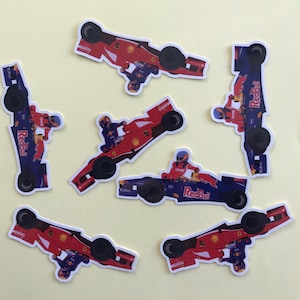 May include: A collection of sticker decals featuring Formula 1 race cars in red and blue. Each sticker depicts a race car with a driver, the Red Bull logo, and other sponsor details. The stickers are arranged on a light yellow background.