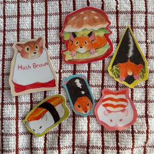 May include: A collection of colorful, illustrated stickers featuring hamsters in various food-themed designs. Includes a hamster in a burger, sushi, and a hash brown. The stickers are on a red and white striped background.
