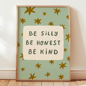 Op de afbeelding: Een rechthoekige wandkunstprint in een houten lijst met een saliegroene achtergrond en verspreide mosterdgele sterren. Een centraal crèmekleurig rechthoekig blok bevat de donkergroene tekst: BE SILLY, BE HONEST, BE KIND.