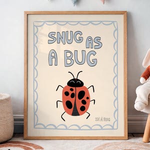 May include: A framed print with the words "SNUG AS A BUG" above a cartoon ladybug illustration. The ladybug is red with black spots and a black head. The print has a light blue border and is in a wooden frame. A stuffed bunny sits on a small stool.