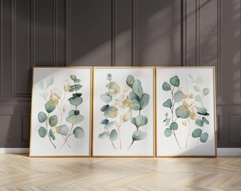 Puede incluir: Tres impresiones artísticas botánicas enmarcadas con hojas de eucalipto en acuarela en tonos verdes y dorados. Las impresiones tienen marcos dorados y se exhiben contra una pared gris oscuro y un suelo de madera clara.