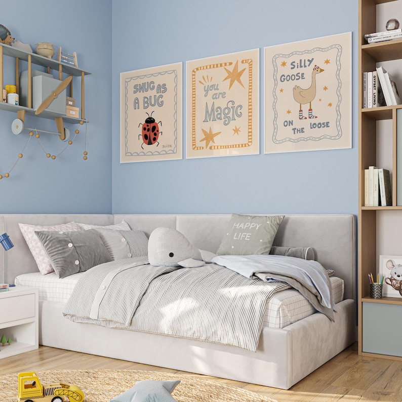 May include: A child's bedroom with a light blue wall, a bed with gray bedding, and three framed prints. The prints feature whimsical illustrations and text, including a ladybug, stars, and a goose. A wooden shelf with a toy airplane is mounted on the wall.