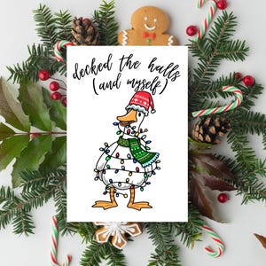 May include: A Christmas card featuring a cartoon duck decorated with Christmas lights, a Santa hat, and a green scarf. The card is surrounded by a wreath of pine branches, candy canes, and gingerbread cookies. The text on the card reads "decked the halls (and myself)".