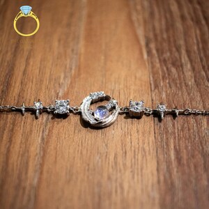 May include: A silver bracelet with a moon and stars design. The bracelet features a large moon charm with a clear stone in the center, surrounded by smaller stars. The bracelet is made of silver and has a delicate chain.