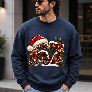 May include: A navy blue crewneck sweatshirt featuring a large graphic of the number 67 in a red and green plaid pattern. The numbers are decorated with a Santa hat, snow, pine cones, holly berries, and glowing string lights.