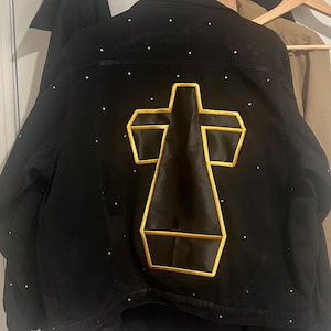 Może przedstawiać: Czarna kurtka dżinsowa z dużym, czarnym satynowym krzyżem w żółtej ramce na plecach. Kurtka jest pokryta małymi, białymi kryształkami.