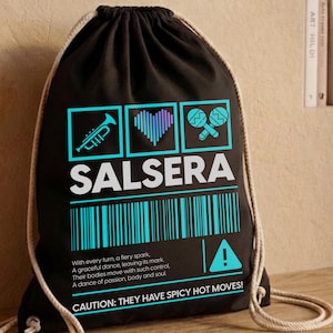 Könnte beinhalten: Ein schwarzer Kordelzugrucksack mit einer Grafik aus Trompete, Herz und Maracas über dem Wort SALSERA in Weiß. Darunter befinden sich ein cyanfarbener Barcode, Text über Tanz und die Warnung: CAUTION: THEY HAVE SPICY HOT MOVES!