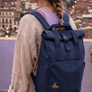 May include: A navy blue roll-top backpack featuring two top handles with a snap button closure, a front zippered pocket, and a small embroidered graphic of the map of Colombia in yellow, blue, and red colors on the lower right corner.